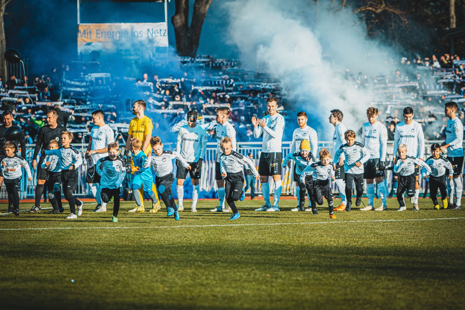 Fußballspieler laufen mit Kindern auf einem Rasenplatz vor jubelnden Fans mit Fahnen und farbigem Rauch ein.